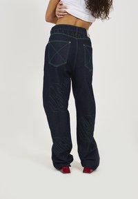 Dark denim wide-leg pants with green stitching, featuring two back pockets and bold embossed logo detailing along the sides.