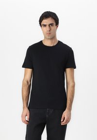Camiseta de algodón negra con mangas cortas, cuello redondo y un diseño ajustado. Llevada con jeans oscuros, contra un fondo blanco liso.