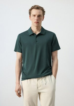 Uomo che indossa una polo a maniche corte verde scuro e pantaloni beige chiaro, in piedi con una mano in tasca su sfondo bianco uniforme.