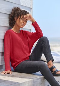 Rotes Langarmoberteil aus leichtem Stoff, kombiniert mit dunklen Slim-Fit-Jeans und schwarzen Sandalen. Lockere Passform mit überschnittenen Schultern, minimalistisches Design.