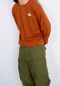 Brązowa koszulka z długim rękawem z białym logo na piersi, w połączeniu z oliwkowymi spodniami cargo z bocznymi kieszeniami i luźnym krojem.