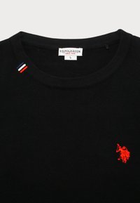 Črna pletena majica s kratkimi rokavi, ki ima rdeč vezen logotip na prsih in tri barvne naglasne črte na rami.
