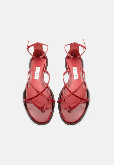 Κόκκινα επίπεδα σανδάλια με λεπτές σταυρωτές λουρίδες και αγκράφες στον αστράγαλο, εμφανίζονται από πάνω σε λευκό φόντο.