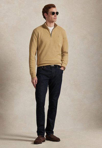 Άνδρας που φοράει μπεζ πουλόβερ με φερμουάρ μέχρι τη μέση, σκούρα τζιν, καφέ μοκασίνια και γυαλιά ηλίου, στέκεται με το ένα χέρι στην τσέπη μπροστά σε απλό φόντο.