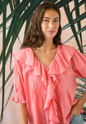 Junge Frau mit langen braunen Haaren, die eine rosa Bluse mit Rüschen und Jeans trägt, steht mit einer Hand in der Hüfte vor einem grünen, blätterreichen Hintergrund.