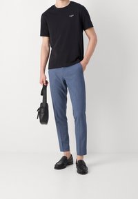 Osoba v černém tričku, modrých slim-fit kalhotách a černých mokasínách, drží malou černou koženou tašku v jedné ruce.