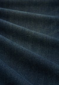 Ciemny materiał dżinsowy o gładkiej fakturze, ukazujący falujące fałdy. Kolor wydaje się głęboko niebieski z subtelnymi odcieniami i klasycznym wzorem tkanym.