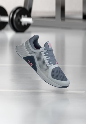 Chaussure de sport grise avec des panneaux en mesh et un logo rose semble flotter dans une salle de sport, avec des poids et équipements flous en arrière-plan.