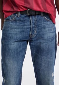 Vaqueros de mezclilla azul con un corte relajado, presentan zonas desgastadas y parches rasgados. Terminado con un cinturón negro y bolsillos frontales.