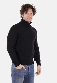 Maglione a collo alto nero con texture a coste e design aderente, abbinato a jeans in denim azzurro chiaro. Il modello si trova su uno sfondo neutro.