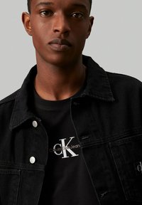 Chaqueta de mezclilla negra con broches plateados, superpuesta a una camiseta negra con el logo y texto blanco de Calvin Klein. Textura suave, diseño clásico.