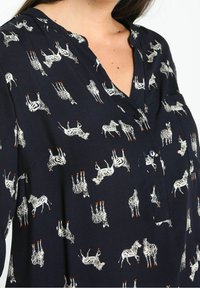 Chemisier bleu marine avec col boutonné, présentant un motif zèbre répétitif en blanc avec des accents orange. Tissu doux avec une coupe décontractée.