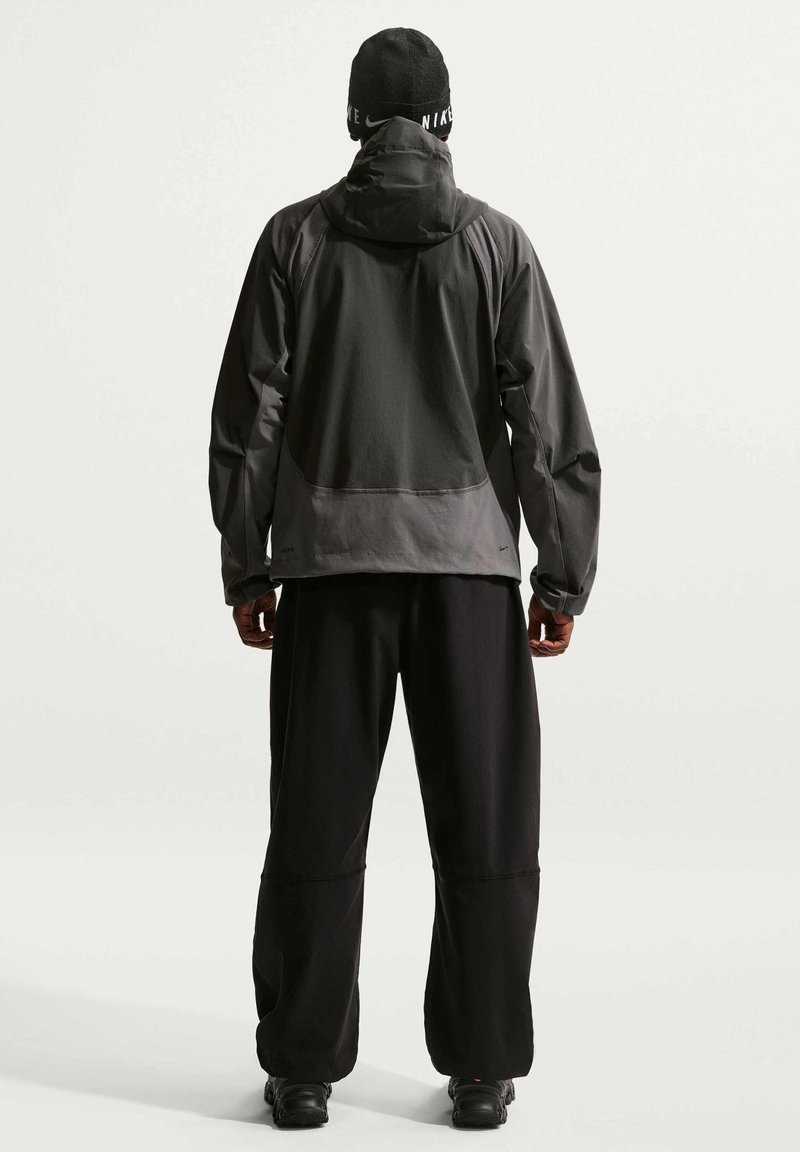 Uomo che indossa giacca Nike nera e grigia, pantaloni neri, scarpe nere e berretto Nike nero, in piedi di spalle contro uno sfondo chiaro e uniforme.