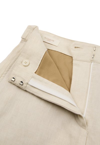 Cintura de pantalones beige mostrando botón, cierres de gancho y ojo, cremallera y etiqueta interior que dice "SLOWEAR INCOTEX."