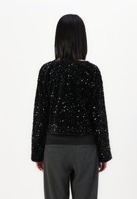 Giacca corta nera con paillettes e maniche lunghe, caratterizzata da una texture morbida e un collo rotondo, abbinata a pantaloni grigi a vita alta.