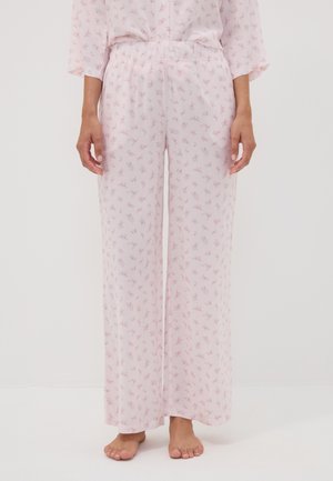 Mujer con pantalones de pijama anchos de color rosa claro con pequeño estampado floral, de pie descalza sobre un suelo blanco.