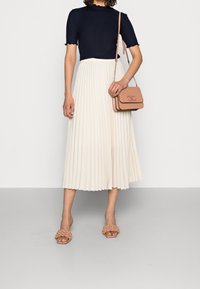 Haut à manches courtes navy, jupe midi plissée crème, sandales à talons nus avec des détails texturés, et un petit sac à main rose avec une bandoulière en chaîne.