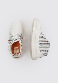 Zapatillas de lona blancas con parte superior de tela estampada que presenta diseños geométricos negros, suela de goma y cordones ajustables con cuentas decorativas.