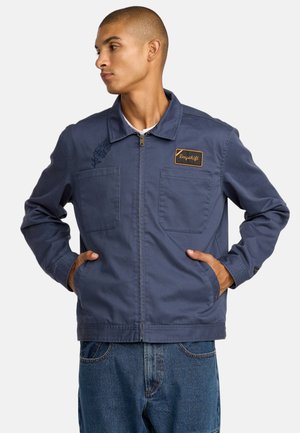 Mann trägt eine marineblaue Arbeitsjacke mit Reißverschluss und einem Aufnäher mit der Aufschrift "Dayshift" und hat die Hände in den Taschen, schaut nach links.