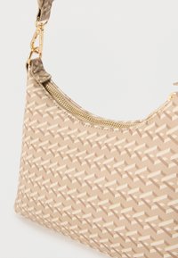 Sac à bandoulière beige à motif avec fermeture éclair dorée et fermoir doré sur fond uni, montrant un gros plan de la texture du tissu et des éléments métalliques.