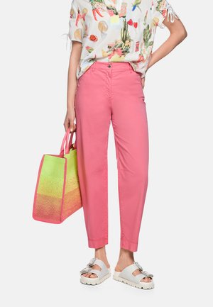 Person in pinker Hose, weißen Sandalen mit großen Schnallen, hält eine bunte Tragetasche, trägt ein weißes Hemd mit mehrfarbigen Drucken.