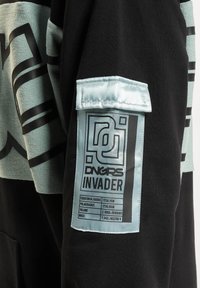 Fekete szövet kapucnis pulóver világoskék grafikai mintával és címkével. A címke geometrikus szöveget és részleteket tartalmaz egy texturált folton.