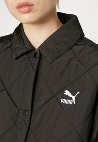 Svart quiltet jakke med krage, med svart trykknapp og hvit Puma-logo på høyre bryst.