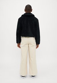 Casaco de fleece preto, cropped, com colarinho, apresentando mangas longas e fechos de correr nos punhos, combinado com calças largas em creme.