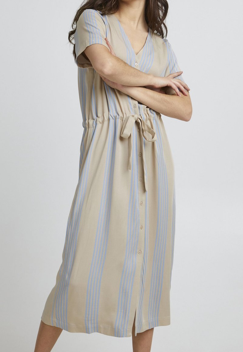 Femme portant une robe beige avec des rayures verticales bleu clair, manches courtes, boutonnée devant et ceinture à la taille, debout avec les bras croisés.