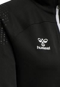 Veste de sport noire avec une texture lisse, ornée d'un logo blanc sur la poitrine et de détails réfléchissants sur l'épaule. Fermeture éclair à l'avant.