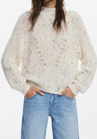 Cremefarbener, gestrickter Pullover mit mehrfarbigen Sprenkeln, rundem Ausschnitt und gerippten Bündchen. Kombiniert mit hellblauen Jeans.