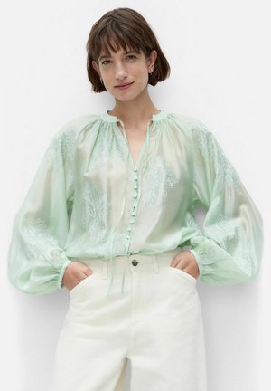 Femme aux cheveux courts bruns portant un chemisier vert menthe transparent avec broderie florale et un pantalon blanc taille haute, mains dans les poches.