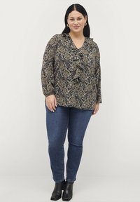 Bloemige blouse met een gerimpelde V-hals, lange mouwen en een donkere achtergrond. Gepaard met blauwe spijkerbroeken en zwarte enkellaarsjes, wat een casual look oplevert.
