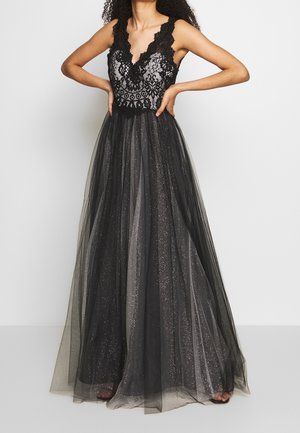 Femme portant une robe noire en dentelle sans manches avec une jupe en tulle scintillant, debout les mains sur les hanches devant un fond uni.