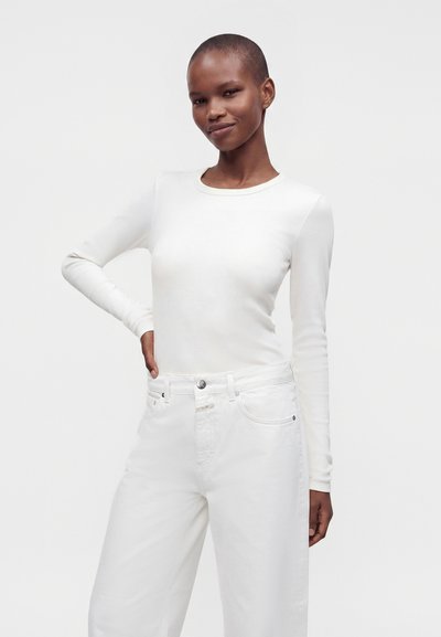 Personne debout aux cheveux courts portant un haut blanc ajusté à manches longues et un pantalon blanc ample sur un fond clair uni.