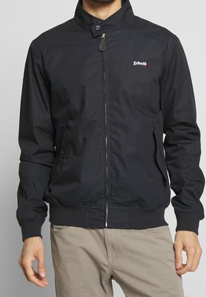 Mann trägt eine schwarze Jacke mit Reißverschluss und Stehkragen sowie beige Hosen, die Hände entspannt an den Seiten, sichtbar vom Kinn bis zur Hüfte.