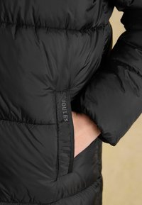 Černá vycpaná bunda s lesklým povrchem, která má boční kapsu a logo "Joules" v kontrastní barvě na rukávu.