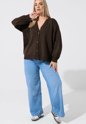 Kvinde iført en mørkebrun cardigan med knapper, lyseblå vidde jeans og hvide sneakers med sorte striber, poserer foran en hvid baggrund.