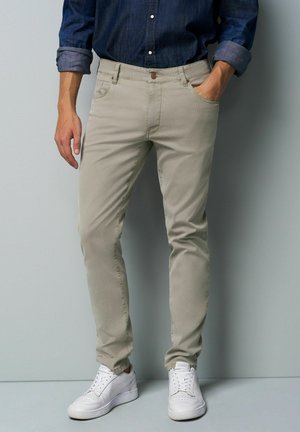 Man wearing beige slim-fit pants, a rolled-up dark blue denim shirt, and white sneakers, standing against a gray wall.