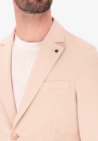 Blazer beige con texture morbida, revers a punta, chiusura a tre bottoni e una tasca sul petto. Indossato sopra una camicia bianca.