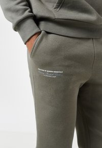 Άτομο με το χέρι στην τσέπη, φορώντας χακί πράσινη φόρμα και φούτερ με λευκό και γκρι κείμενο στο μηρό που γράφει "Δημιουργοί Σύγχρονων Απαραίτητων."