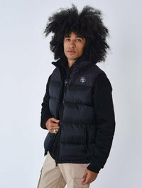 Chaleco de plumas negro con cremallera frontal, que presenta una textura suave y un diseño ajustado. Lleva encima un hoodie negro, complementado con pantalones beige.