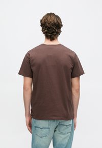 Καφέ βαμβακερό μπλουζάκι με στρογγυλή λαιμόκοψη, κοντά μανίκια και ίσια τελείωμα. Φοριέται με ανοιχτόχρωμο τζιν παντελόνι με τσέπες στο πίσω μέρος.