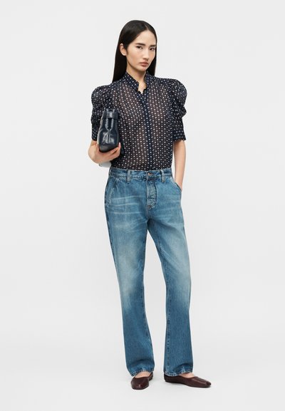 Γυναίκα με διάφανη μαύρη μπλούζα με φουσκωτά μανίκια, φαρδιά μπλε τζιν, καφέ μπαλαρίνες, κρατώντας μικρή μαύρη τσάντα, στέκεται μπροστά σε απλό φόντο.