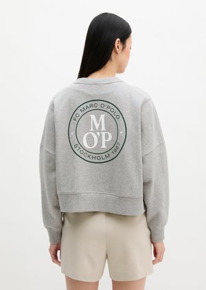 Femme aux longs cheveux foncés portant un sweat-shirt gris avec le logo « FC Marc O'Polo Stockholm 1967 » dans le dos et un short beige.