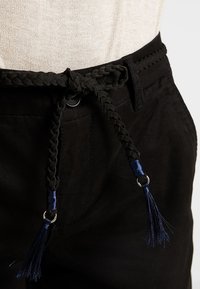 Un pantalon noir doté d'une ceinture tressée avec des pompons bleu marine, fabriqué dans un matériau lisse. Comprend une fermeture à bouton et un détail de couture latérale.