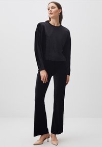 Schwarzes, cropped Langarmoberteil aus strukturiertem Stoff, kombiniert mit schwarzen, weit geschnittenen Hosen. Modell trägt beigefarbene, spitz zulaufende Absätze. Einfaches Design.