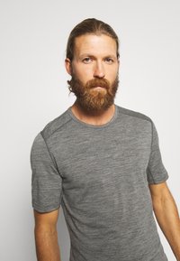 Hombre con barba y cabello largo recogido, vestido con una camiseta gris de manga corta y cuello redondo, posando frente a un fondo liso y claro.
