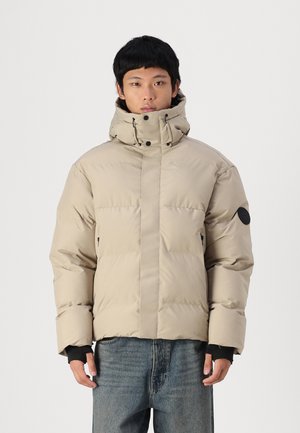 Beige gewatteerde jas met hoge kraag, verstelbare trekkoorden en zwarte accentlabel. Heeft een gemêleerd, gewatteerd ontwerp en zijzakken.