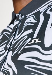 Polo com padrão em preto e branco, feito de um tecido texturizado, apresentando desenhos em espiral, um colarinho escuro e um pequeno logotipo branco no peito.